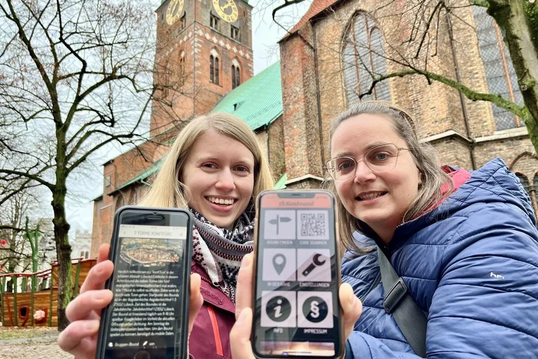 Zwei Frauen stehen vor einer Kirche und halten Smartphones in die Kamera. 