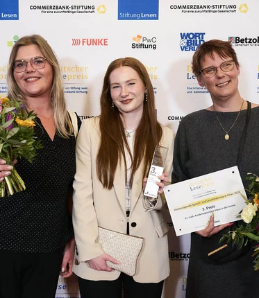 Drei Frauen stehen vor einer Wand und halten Blumen und eine Urkunde.