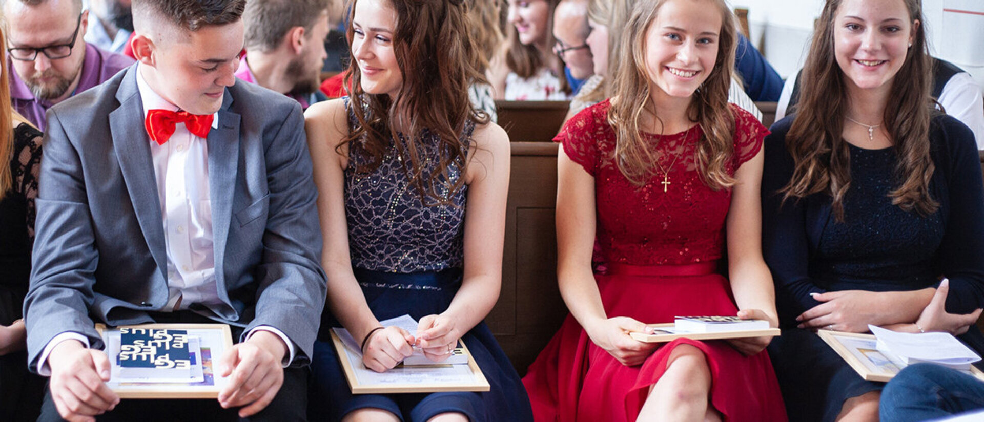 Ein Teenagerjunge mit blau-grauem Sakko, weißem Hemd und roter Fliege sitzt neben drei Teenager-Mädchen. Die Mädchen haben alle lange Haare und sie tragen festliche Kleider. Sie sitzen auf Holzbänken in einer Kirche. Hinter Ihnen sitzen noch mehr Leute auf Holzbänken in mehreren Reihen