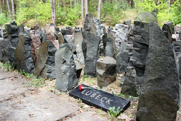 graue Steine stehe aufrecht lose nebeneinander. Eine Platte mit der Aufschrift Lübeck. Darauf liege eine rote Blume. Im Hintergrund Wald.