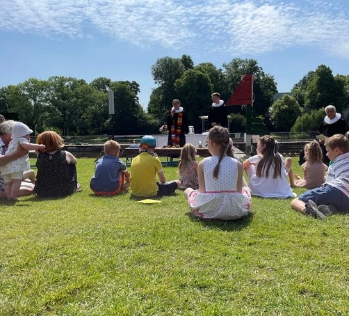 Das Tauffest 2023 im Altstadtbad Krähenteich. Vorne sind Kinder mit ihren Familien sitzend, im Hintergrund Pastorinnen und Pastoren zu sehen. 
