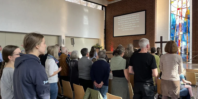 Es stehen mehrere Menschen in Stuhlreihen mit dem Rücken zur Kamera, Sie schauen auf eine Leinwand mit Text. Ein buntes Kirchenfenster und ein Holzkreuz sind daneben zu sehen.
