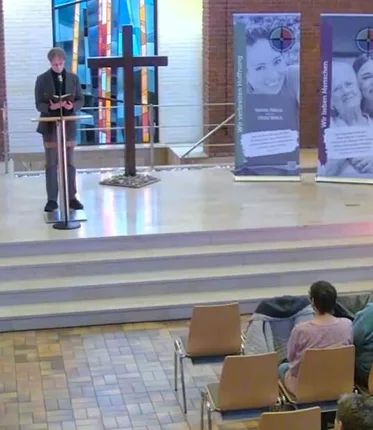 Ein Mann steht in einer Kirche vor einem Pult, vor ihm sitzen Personen auf Stühlen