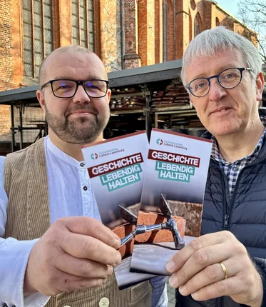 Zwei Männer stehen vor einer Kirche und halten einen Flyer in die Kamera
