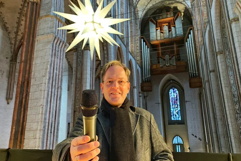 Mann steht in einer Kirche vor einem Leuchtstern und hält lächelnd ein Mikrofon in die Kamera