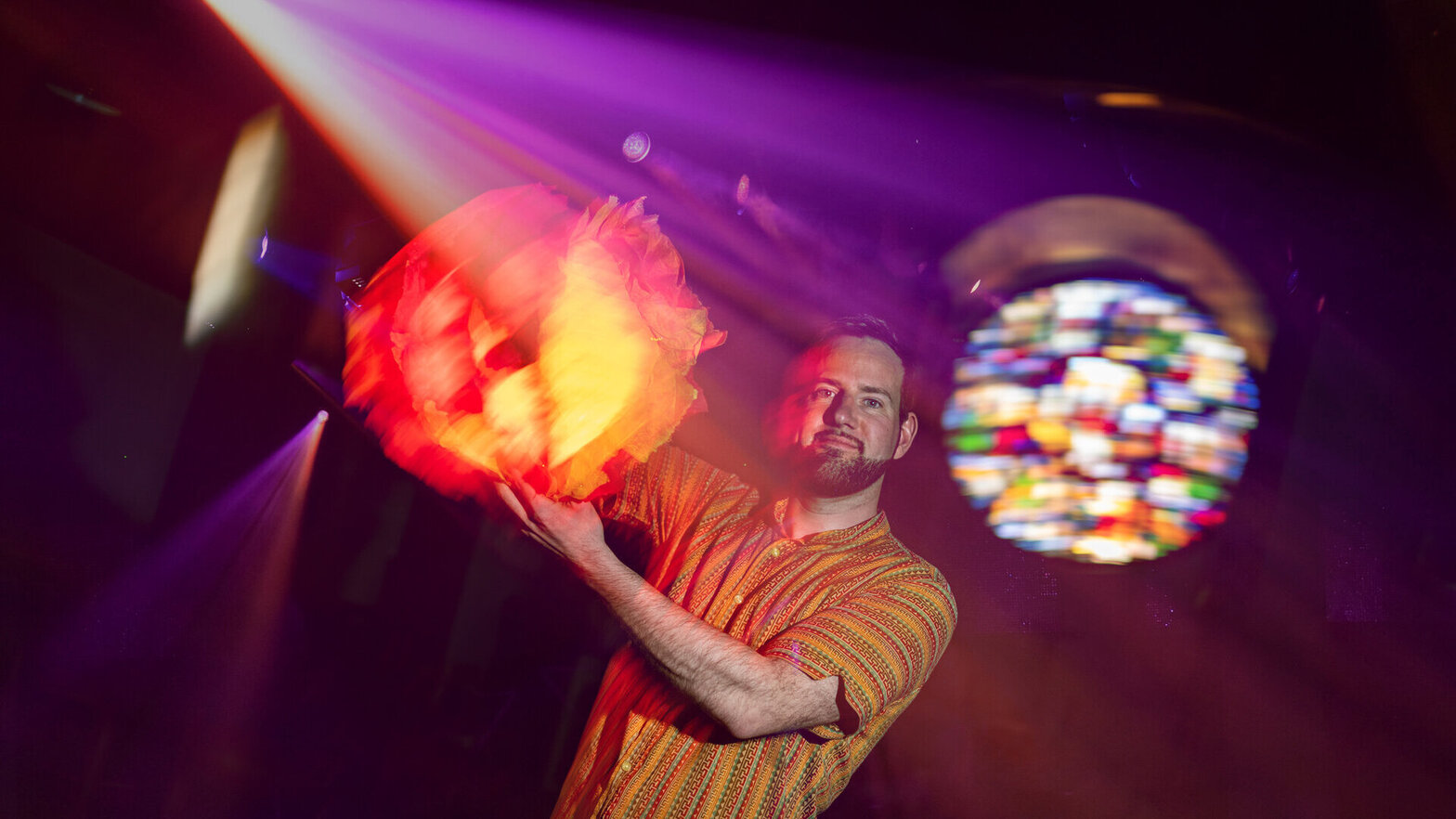 Pastor Christopher Paulsen steht in der dunklen Kirche St. Thomas. Er trägt ein gemustertes Hemd und hält eine runde orange-farbene Laterne in die Höhe. Violette und rote Lichtstrahlen durchziehen die Szene. Im Hintergrund ist das erleuchtete Kirchenfenster zu sehen.