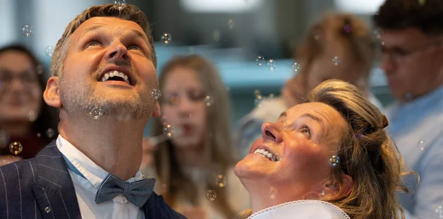 Ein Mann und eine Frau schauen strahlend nach oben. Man sieht undeutlich im Hintergrund eine weibliche Person mit langen Haaren, die Seifenblasen macht.