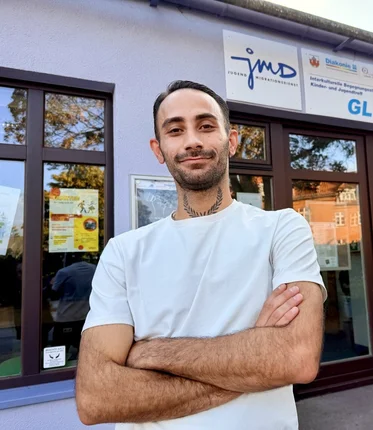 Ein Mann mit weißem T-Shirt und dunklen kurzen Haaren und Drei-Tage-Bart steht vor einem Gebäude mit großen Türen und Fenstern. Er hat die Arme vor sich verschränkt.
