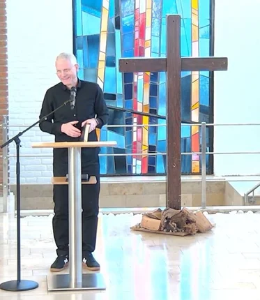 Ein Mann mit Brille und grauen Haaren steht hinter einem Stehpult und hält ein Buch. Im Hintergrund steht ein Holzkreuz und ein buntes Kirchenfenster ist zu sehen