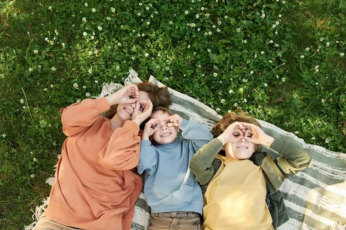 Draufsicht auf eine Frau und zwei Jungen, die zusammen auf einer Picknickdecke liegen und lächelnde Gesichter machen - sie halten die Finger zu einer Brille an die Augen.