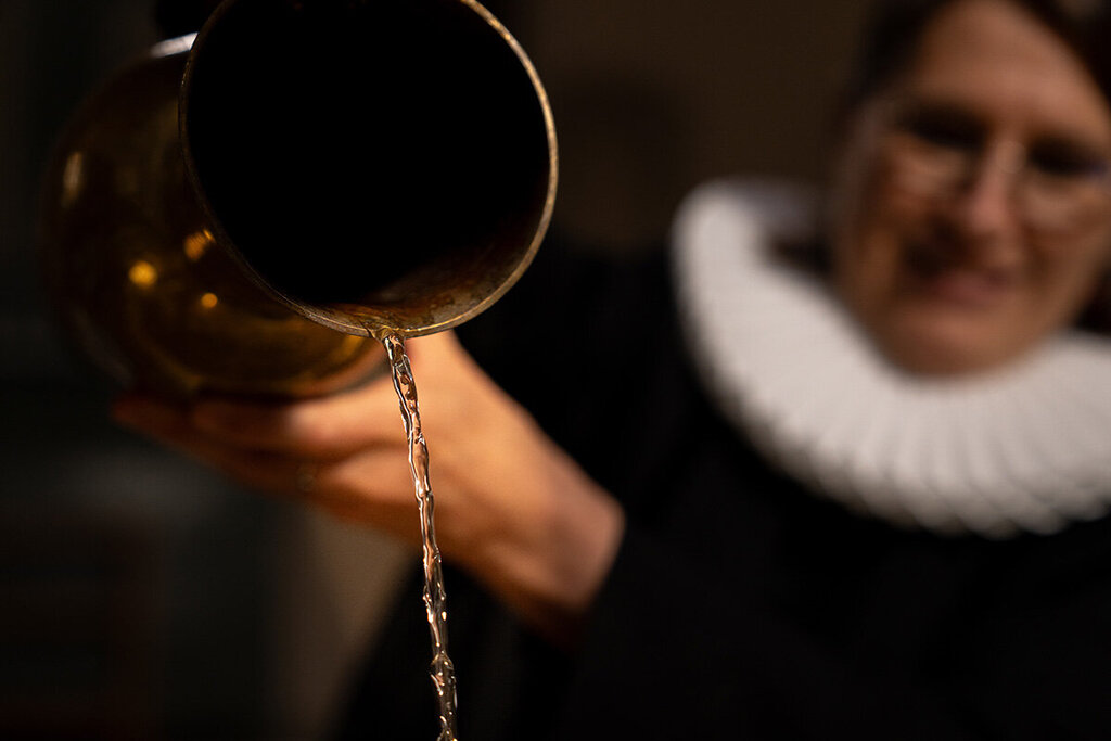 Eine Frau im schwarzen Talar und weißer Halskrause ist nur verschwommen zu sehen. Sie hält ein goldenes Gefäß aus dem Wasser fließt; das Gefäß und das Wasser sind scharf zu sehen. Die Frau trägt eine Brille.