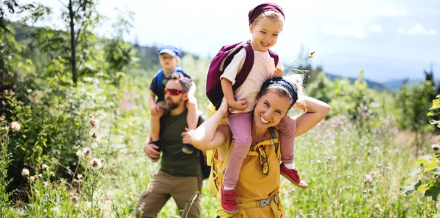 Ein Mann und eine Frau tragen jeweils ein Kind auf Ihrer Schulter und gehen in der Natur spazieren