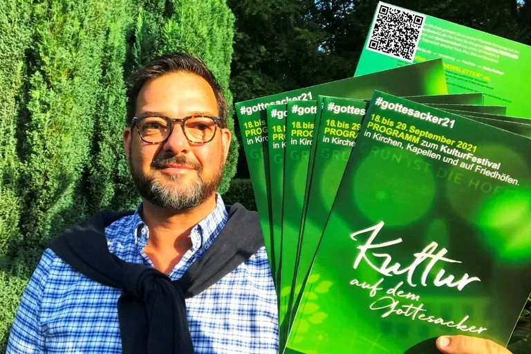 Ein Mann mit Brille hält Flyer in die Kamera. Sie sind grün. 