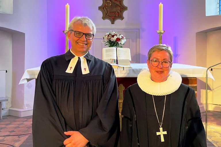Zwei Menschen in Talaren. Im Hintergrund der Altar einer Kirche.