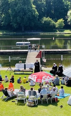 Blick von oben in das Altstadtbad Krähenteich im Jahr 2023. Es sind viele Familien zu sehen, die im Sonnenschein sitzen und den Gottesdienst mit den Pastor:innen mitmachen.