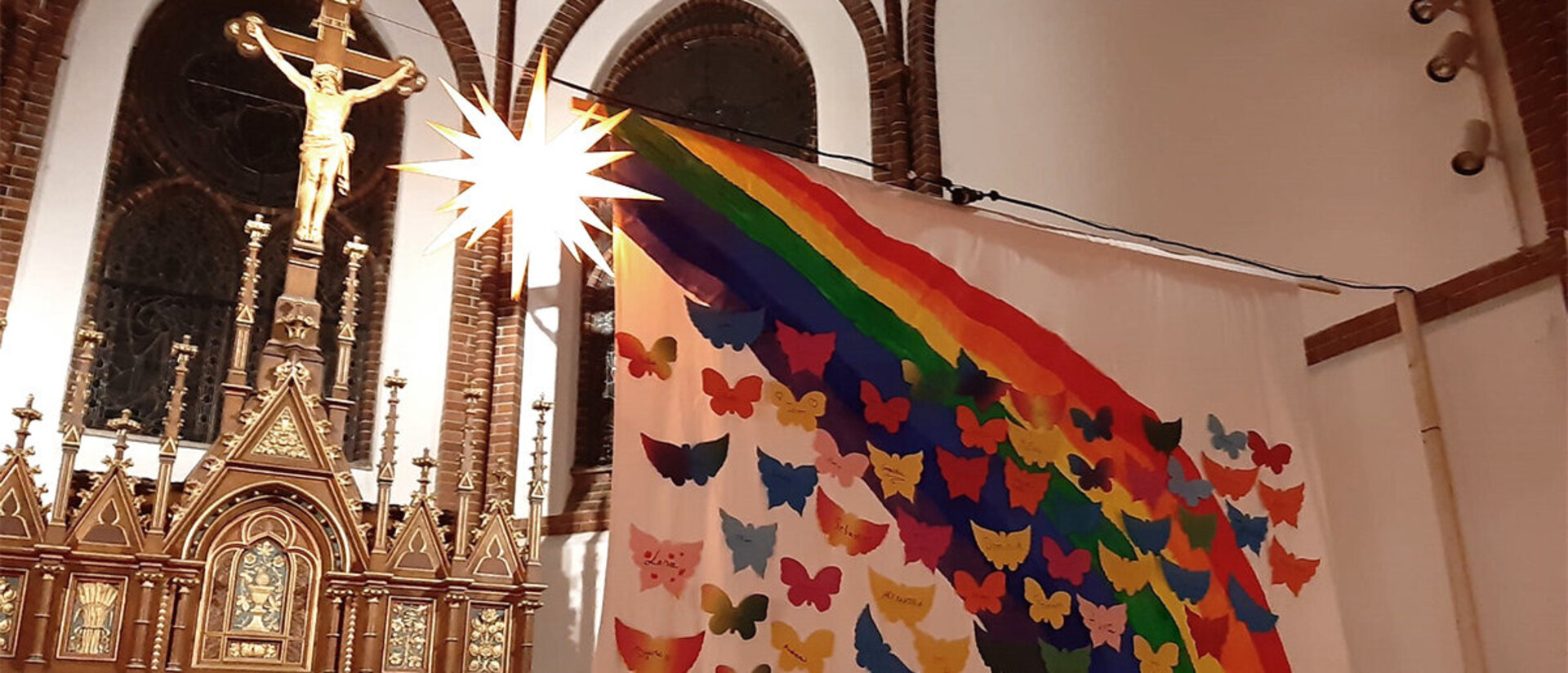 Ein Altar mit Jesus am Kreuz im Hintergrund, einem hell strahlenden gelben Stern und im Vordergrund eine Leinwand mit einem bunten Regenbogen und vielen bunten kleinen Schmetterlingen, welche gen Himmel, nach oben fliegen