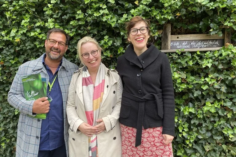 Drei Personen stehen lächelnd vor einer mit Efeu bewachsenen Backsteinmauer.