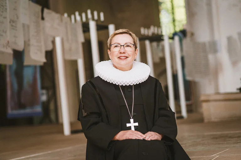 Frau im Talar sitzt lächelnd auf einer Treppenstufe in einer Kirche 