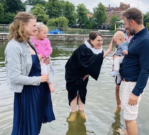 Nach Töchterchen Eva (vor zwei Jahren) lassen Marlene und Tobias Reinberger Sohnemann Johan von Pastorin Margrit Wegner taufen. Sie alle stehen im Wasser, die Pastorin lächelt und tauft Johan.