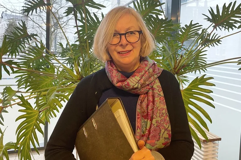 Eine Frau steht vor einer Pflanze und trägt einen Aktenordner unter dem Arm.