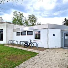 Ein flaches weißes Gebäude mit Eingang auf der rechten Seite, im Vordergrund ein Steinweg und Rasenfläche