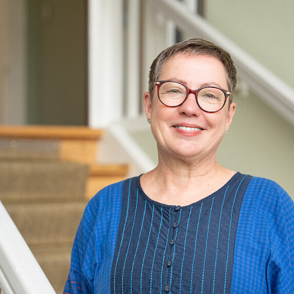 Eine Frau mit blonden Haaren und Brille. Sie schaut lächelnd in die Kamera. Sie trägt ein blaues Oberteil.