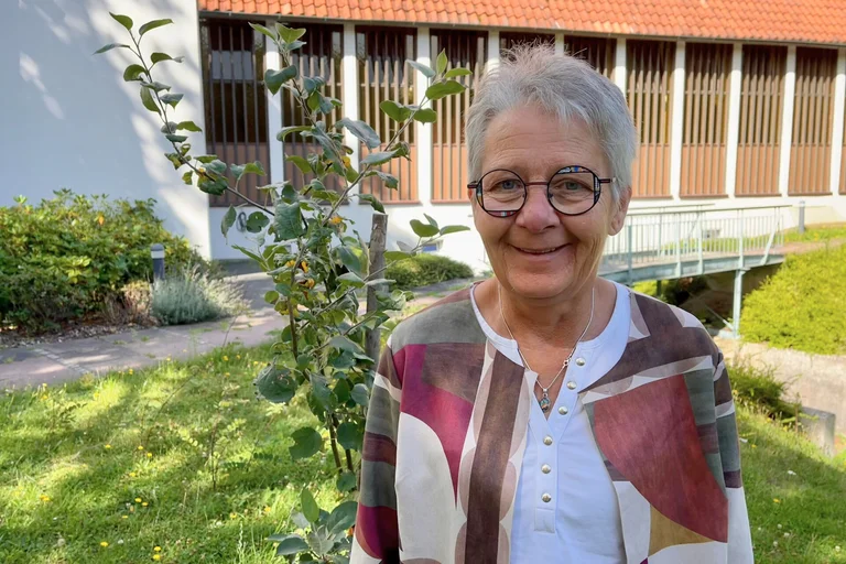 Frau steht vor einem kleinen Apfelbaum und lächelt.