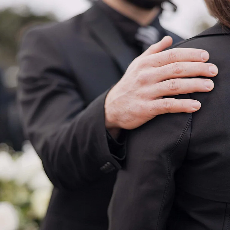 Im Hintergrund verschwommen sieht man einen weißen Blumenstrauß und eine Person mit dunklem Anzug. Im Vordergrund scharf abgebildet hält eine Mann, dessen Kopf man nicht sehen kann, seine rechte Hand tröstend auf der Schulter einer Frau in schwarzer Kleidung und dunklen Haaren. Auch ihr Gesicht kann man nicht sehen. Der Mann trägt ebenfalls schwarze Kleidung: Ein schwarzes Sakko und ein dunkles Hemd mit dunkler Krawatte.