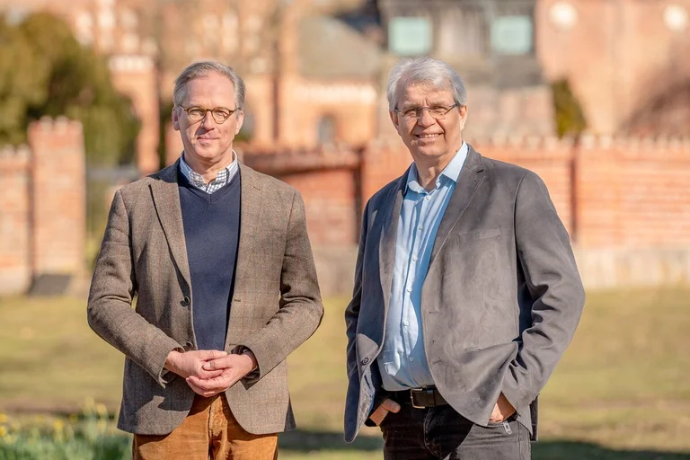 Zwei Männer stehen draußen nebeneinander. Im Hintergrund unscharf ein Gebäudeteil aus rotem Backstein.