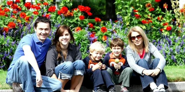 Vater, Teenagertochter, blonder Sohn, braunhaariger Sohn und Mutter mit Sonnenbrille sitzen auf einem Kantstein vor einem Blumenbeet.