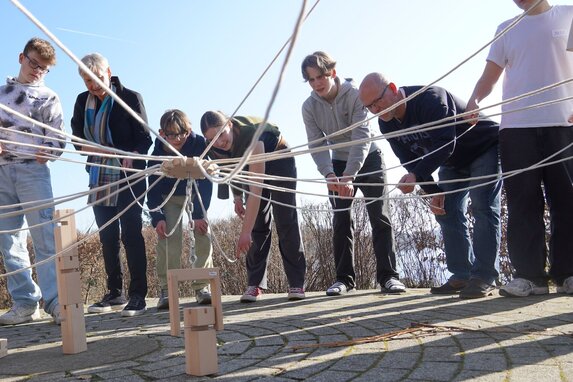 Menschen stehen im Kreis und halten Seile, die in der Mitte zusammenlaufen.