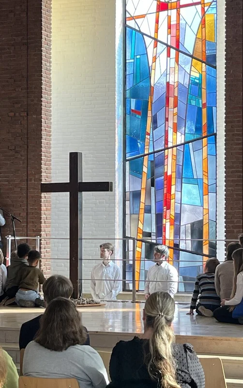 Mehrere Menschen in einer Kirche-2 Personen stehen hinter einem Kreuz in einem Taufbecken, andere stehen darum herum und einige auf Stühlen vor ihnen