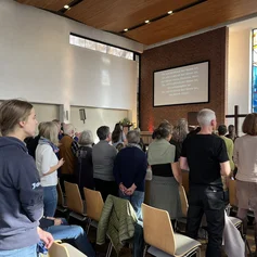 Es stehen mehrere Menschen in Stuhlreihen mit dem Rücken zur Kamera, Sie schauen auf eine Leinwand mit Text. Ein buntes Kirchenfenster und ein Holzkreuz sind daneben zu sehen.