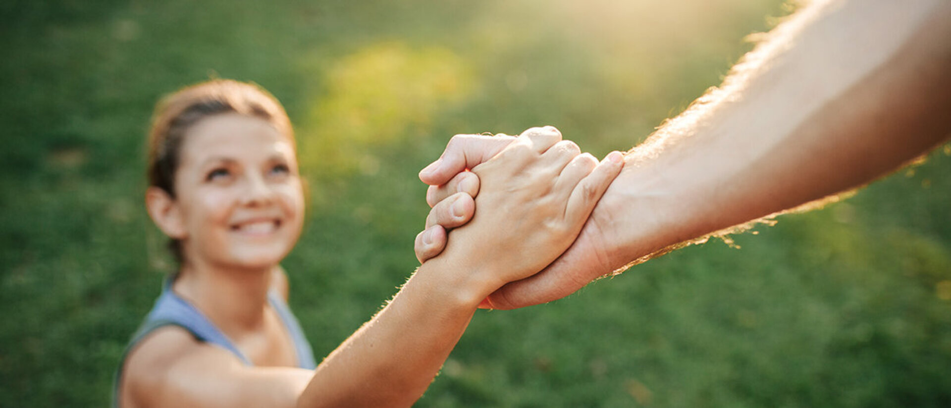 Eine Frau sitzt auf dem Rasen und hält die Hand einer Person von der nicht mehr zu sehen ist. Es scheint die Sonne, die man aber nicht sieht. Die Frau lächelt.