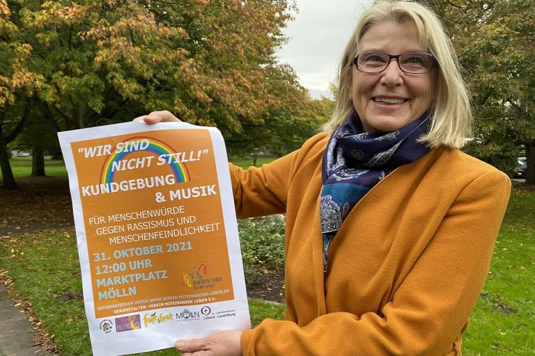 Frau steht mit einem Plakat lächelnd vor herbstlichen Bäumen und schaut in die Kamera. 