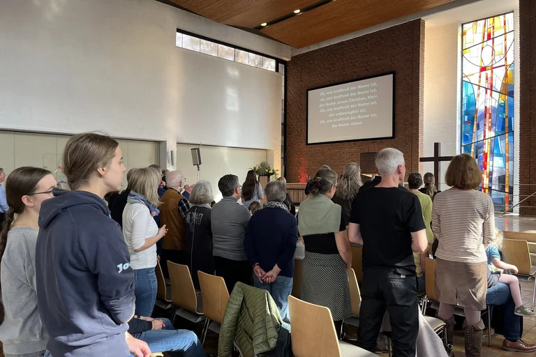 Es stehen mehrere Menschen in Stuhlreihen mit dem Rücken zur Kamera, Sie schauen auf eine Leinwand mit Text. Ein buntes Kirchenfenster und ein Holzkreuz sind daneben zu sehen.