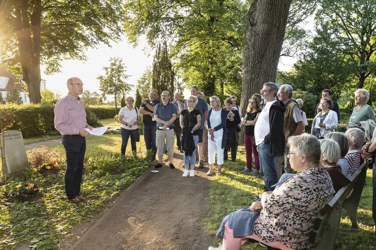 Ein Mann steht vor einem Grab im Abendlicht und spricht zu einer Gruppe Menschen