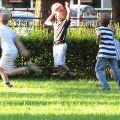 Vier Jungen spielen auf einem Rasen der Teils sonnenbeschienen ist. Der zweite Junge von links hält einen Ball über seinem Kopf