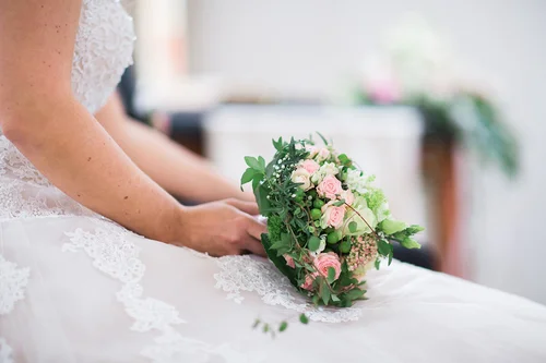 Eine Braut, dessen Kopf nicht zu sehen ist, sitzt auf einem Stuhl. Sie trägt ein kurzärmeliges weißes Kleid und hält einen Brautstrauß rosa-grün-farben vor sich auf dem Schoß.
