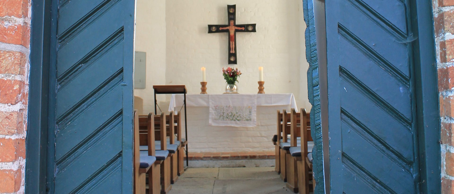 Eine offene blaue Kirchentür gibt den Blick frei zu einem Kirchenaltar auf dem zwei große, weiße, brennende Kerzen zu sehen sind. Zwischen den Kerzen steht eine runde Glasvase mit Blumen, darüber hängt ein Altarkreuz. Im vordergrund sind braune Holz-Kirchenbänke mit blauen Sitzauflagen zu sehen.