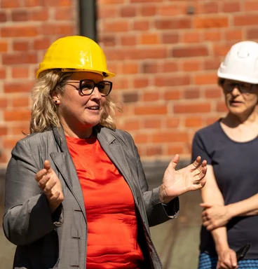 Eine Frau mit rotem Oberteil und gelbem Oberteil spricht vor einer Backsteinmauer