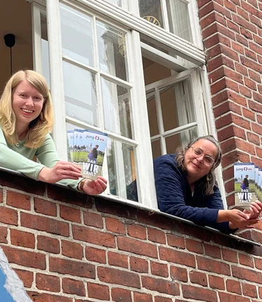 Zwei Frauen schauen aus einem Fenster und halten Flyer in den Händen.