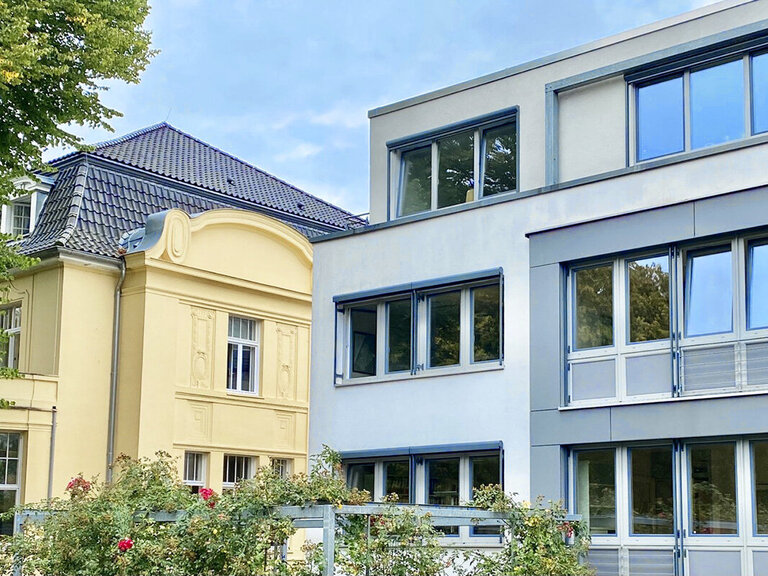 Alt- und Neubau der Kirchenkanzlei - Auf der linken Seite der Altbau mit gelber Fassade und dunklem Dach, rechts der Neubau in Grautönen und Flachdach