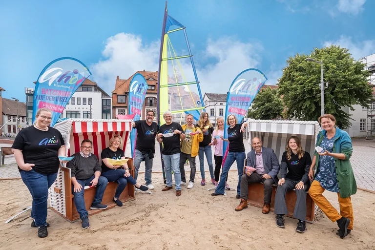 Mehrere Menschen auf einer Sandfläche mit Strandkörben lächeln in die Kamera.