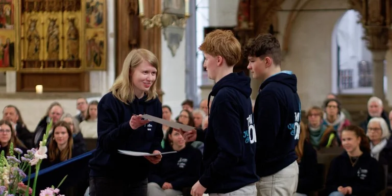 Eine blonde Frau überreicht zwei Jugendlichen eine Urkunde