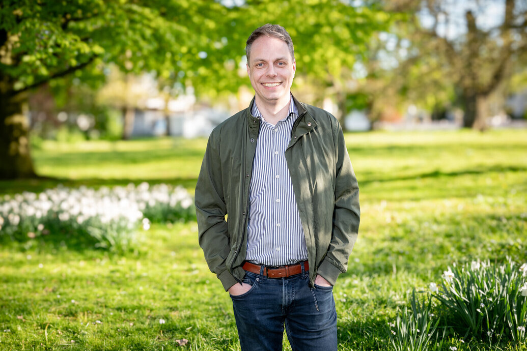  Ein Mann lächelt in die Kamera. Er trägt eine dunkelblaue Jeanshose mit braunem Gürtel, ein helles Hemd und eine dunkelgrüne Jacke. Im Hintergrund ist eine Wiese mit Frühlingsblumen im Sonnenschein zu sehen und eine paar Zweige mit hellgrünen Blättern ragen von links oben ins Bild.
