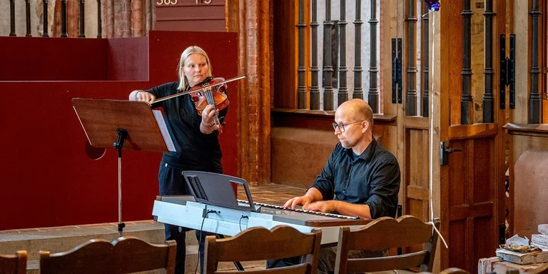 Ein Mann am Klavier und eine Frau mit Geige spielen in einer Kirche. 