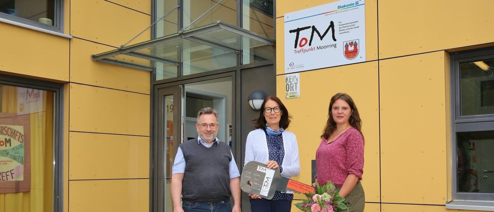 Ein Mann und zwei Frauen stehen vor einem gelben Gebäude und halten einen großen Schlüssel aus Pappe.