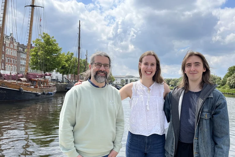Eine Frau und zwei Männer stehen am Wasser mit einem Segelschiff im Hintergrund.