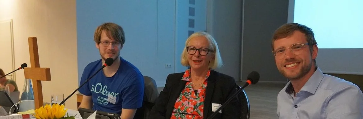 Eine Frau und zwei Männer sitzen an einem Tisch mit Mikrofonen und einem Holzkreuz.